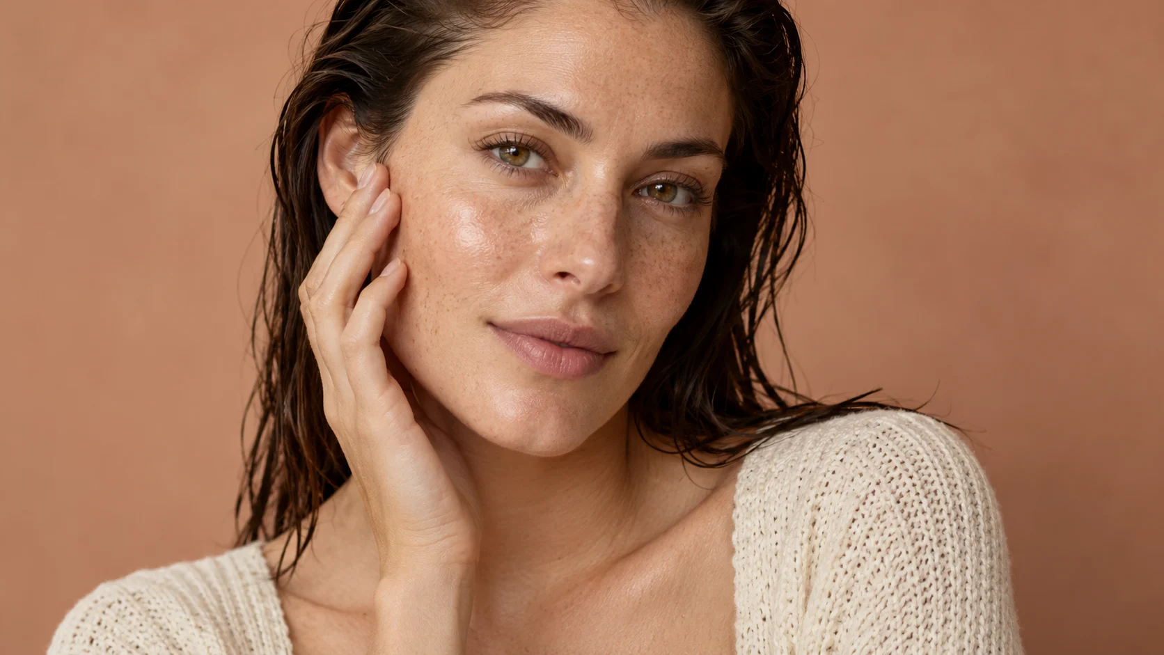 An adult woman with freckles touching her cheek against a warm tan backdrop.